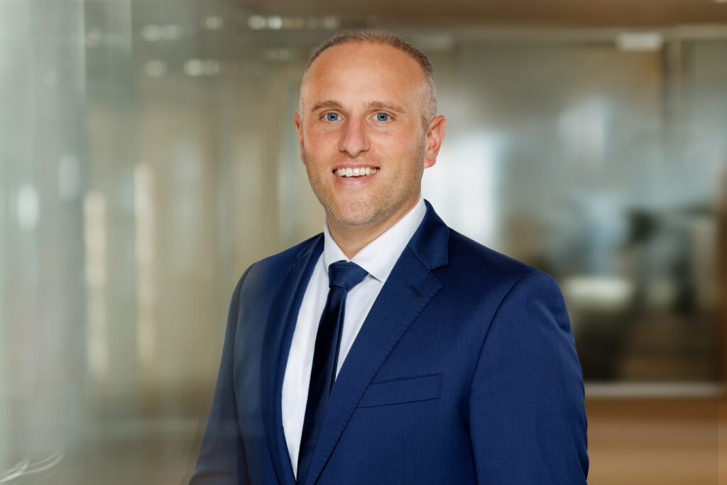 Porträt von Florian Henke, Gewerbeimmobilien, Projektentwicklung für die Regionen Göppingen, Biberach und Heidenheim, im Business-Outfit, Dunkelblauer Anzug, Dunkelblaue Krawatte und weißes Hemd, vor einem modernen Bürohintergrund.