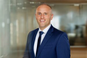 Porträt von Florian Henke, Gewerbeimmobilien, Projektentwicklung für die Regionen Göppingen, Biberach und Heidenheim, im Business-Outfit, Dunkelblauer Anzug, Dunkelblaue Krawatte und weißes Hemd, vor einem modernen Bürohintergrund.