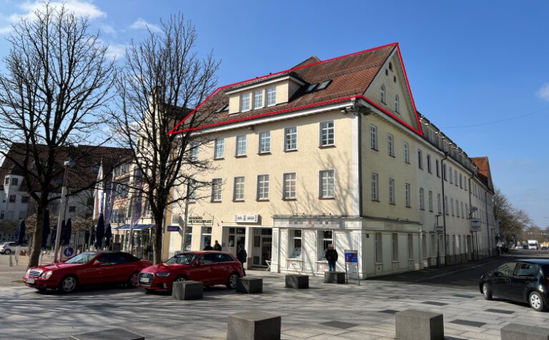 Hellgelbes Geschäftshaus am Göppinger Schillerplatz, blauer Himmel, parkende Autos im Vordergrund, Menschen am Vorbeigehen und Bäume ohne Blätter. Der oberste Stock ist umrandet, dies ist die Mieteinheit.