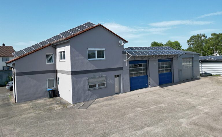 Werkstattgebäude mit grauer Fassade, 3 Rolltore, Büroanbau links und großes befestigtes Grundstück im Vordergrund.