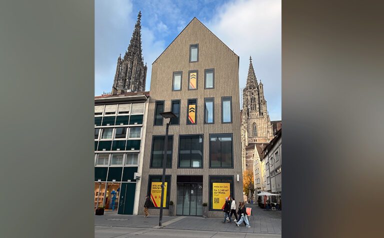 Ein modernes, mehrstöckiges Gebäude mit spitzem Giebel steht in einer Einkaufsstraße in Ulm. Im Hintergrund sind die Türme des Ulmer Münsters zu sehen. Vor dem Gebäude gehen einige Passanten vorbei, und im Erdgeschoss hängen gelbe Plakate der fgi mit schwarzer und roter Schrift in den Fenstern.