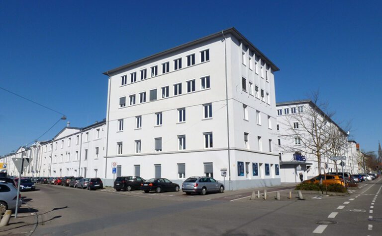 Außenansicht des sanierten Büro- und Gewerbegebäudes Röhrenwerk in Ulm mit weißer Fassade, mehreren Etagen und parkenden Autos entlang der Straße bei blauem Himmel.