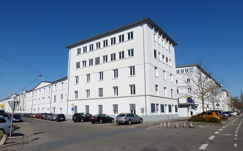 Außenansicht des sanierten Büro- und Gewerbegebäudes Röhrenwerk in Ulm mit weißer Fassade, mehreren Etagen und parkenden Autos entlang der Straße bei blauem Himmel.