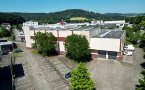 Luftaufnahme eines großflächigen Gewerbeareals mit Produktions- und Lagerhallen, befestigten Hofflächen und Parkplätzen in gewerblicher Lage, umgeben von Grünflächen und Hügellandschaft.