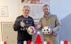 2 Herren lachen in die Kamera, an der einen Hand symbolisch Daumen nach oben in der anderen Hand jeweils ein Fußball. Es war die Spendenübergabe von fgi, stellvertretend der Geschäftsführer Sebastian Fercho, an das Rupert-Mayer-Haus, stellvertretend Herr Michael Hoyler. Im Hintergrund ist eine grau, weiße Wand und ein Plakat des Rupert-Mayer-Hauses zu sehen. Als dekoratives Element ist am unteren Rand eine Grafik mit Geschenken und Weihnachtsbaum in Rot, Weiß und Beige.