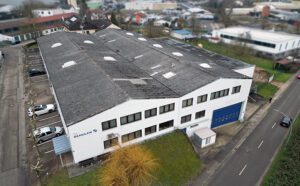 Luftaufnahme einer großen Produktionshalle in Gemmingen mit weißer Fassade, blauem Rolltor und angrenzenden Parkplätzen in einem Gewerbegebiet. Im Vordergrund ist eine Straße, im Hintergrund sind unscharf weitere FIrmengebäude zu sehen.