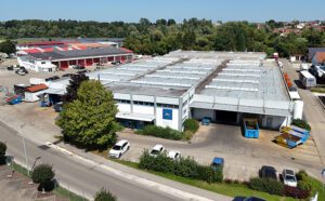 Großes Produktionsreal in Babenhausen. Flachdach, mehrere Hallenteile. Eingang und Tore sind sichtbar. Im Vordergrund eine befestigte Außenfläche mit Parkplätzen umsäumt von Büschen und Bäumen. Davor ist eine Straße und im Hintergrund weitere Gewerbebetriebe.